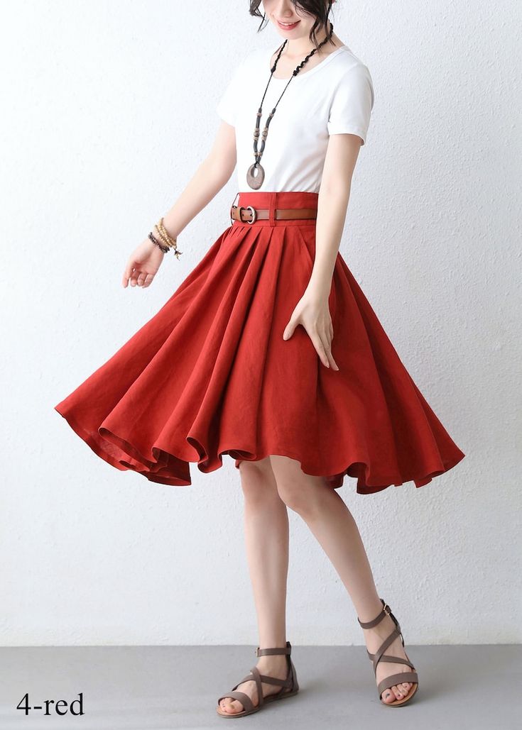 a woman wearing a white tshirt and red skirt with a brown belt, sandals, a necklace, and a bracelet stands against a white wall. the skirt is flared and appears to be made of linen or a similar lightweight fabric. the woman is positioned at an angle, with the skirt twirling slightly.