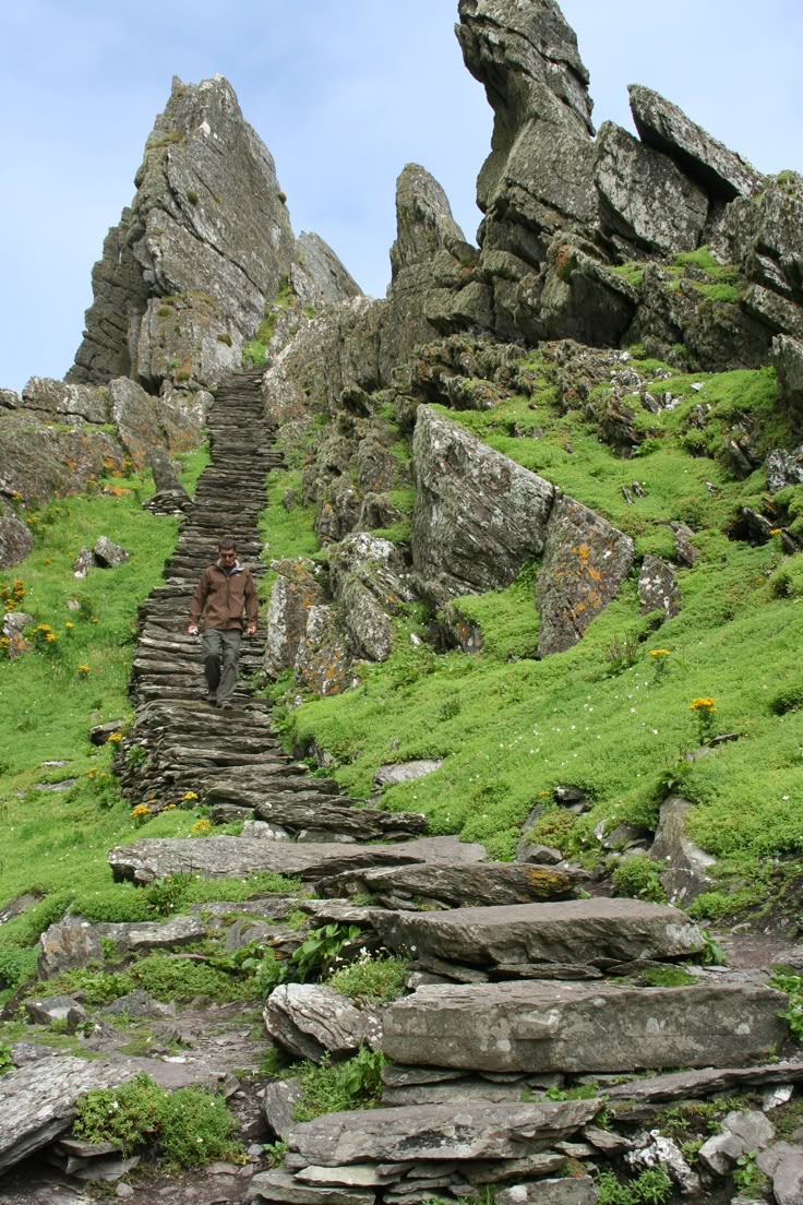 a man walks down a steep, stone staircase on skellig michael in ireland. the staircase winds down through a rocky, mountainous landscape covered in green grass and moss. large, jagged rocks are prominent features of the scenery. the sky is overcast.