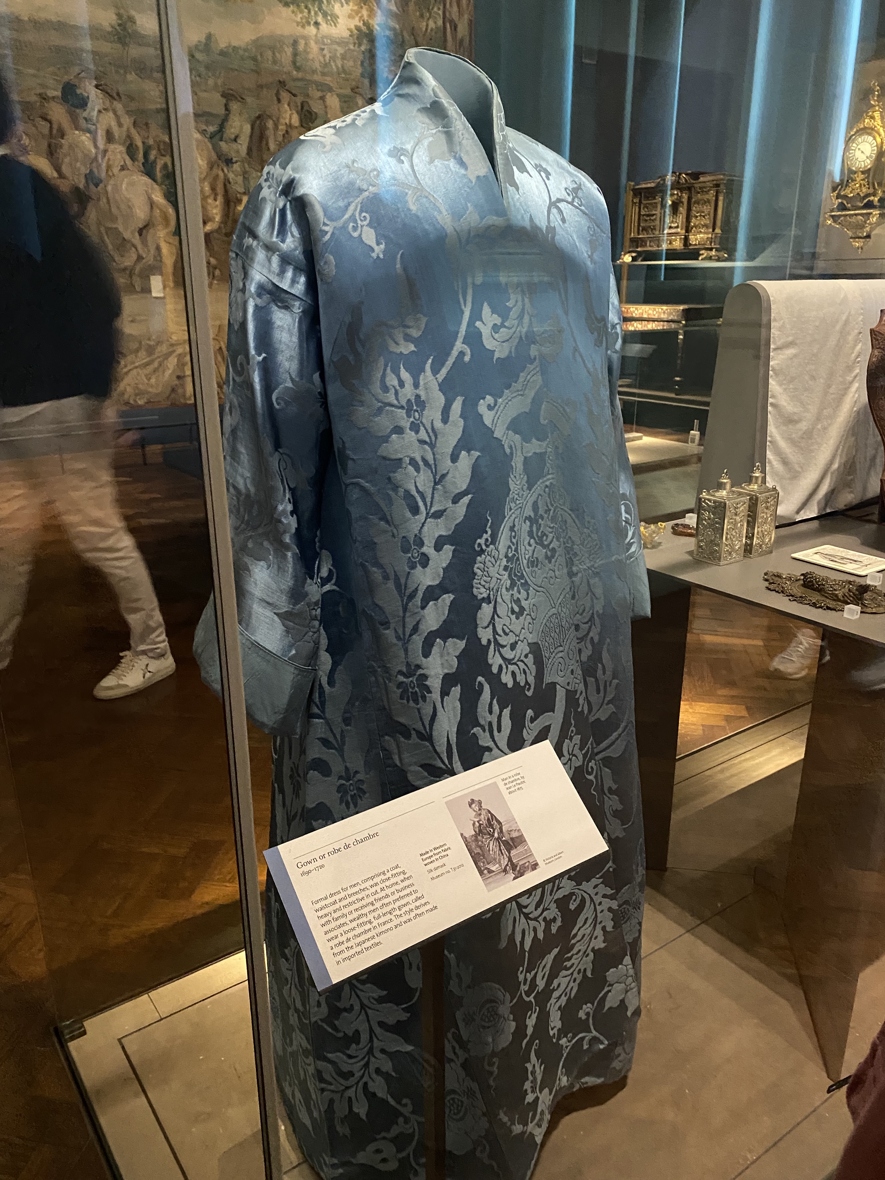 a longsleeved, blue damask gown or robe is displayed in a glass case at a museum. a descriptive placard stands beside the gown. in the background, there are other artifacts and a tapestry, and the reflection of museum visitors can be seen in the glass case.