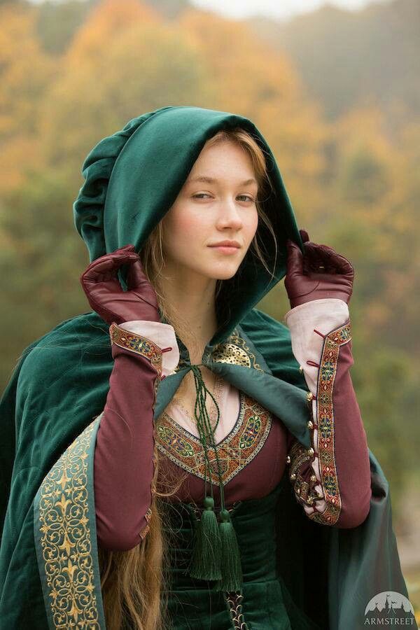 a young woman in a medievalstyle green hooded cloak with a brown dress. she is wearing leather gloves with embroidered cuffs. the dress has a decorative neckline with tassels. she is standing outdoors with a blurred background of trees. the image has a fantasy or historical theme.