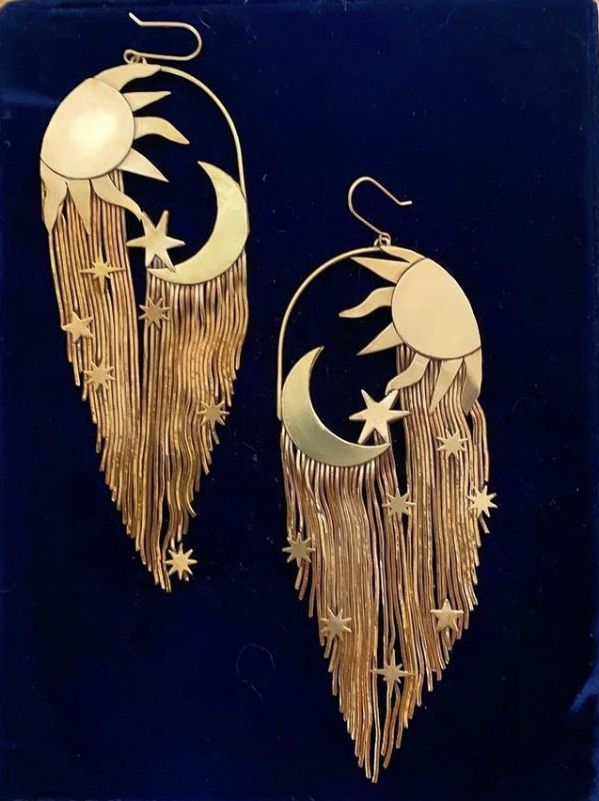 the image shows a pair of ornate goldtoned earrings against a dark blue background. each earring features a sun at the top with rays extending outwards, a crescent moon shape, and several star charms scattered among a fringe of thin golden chains. the earrings have a hook for piercing.