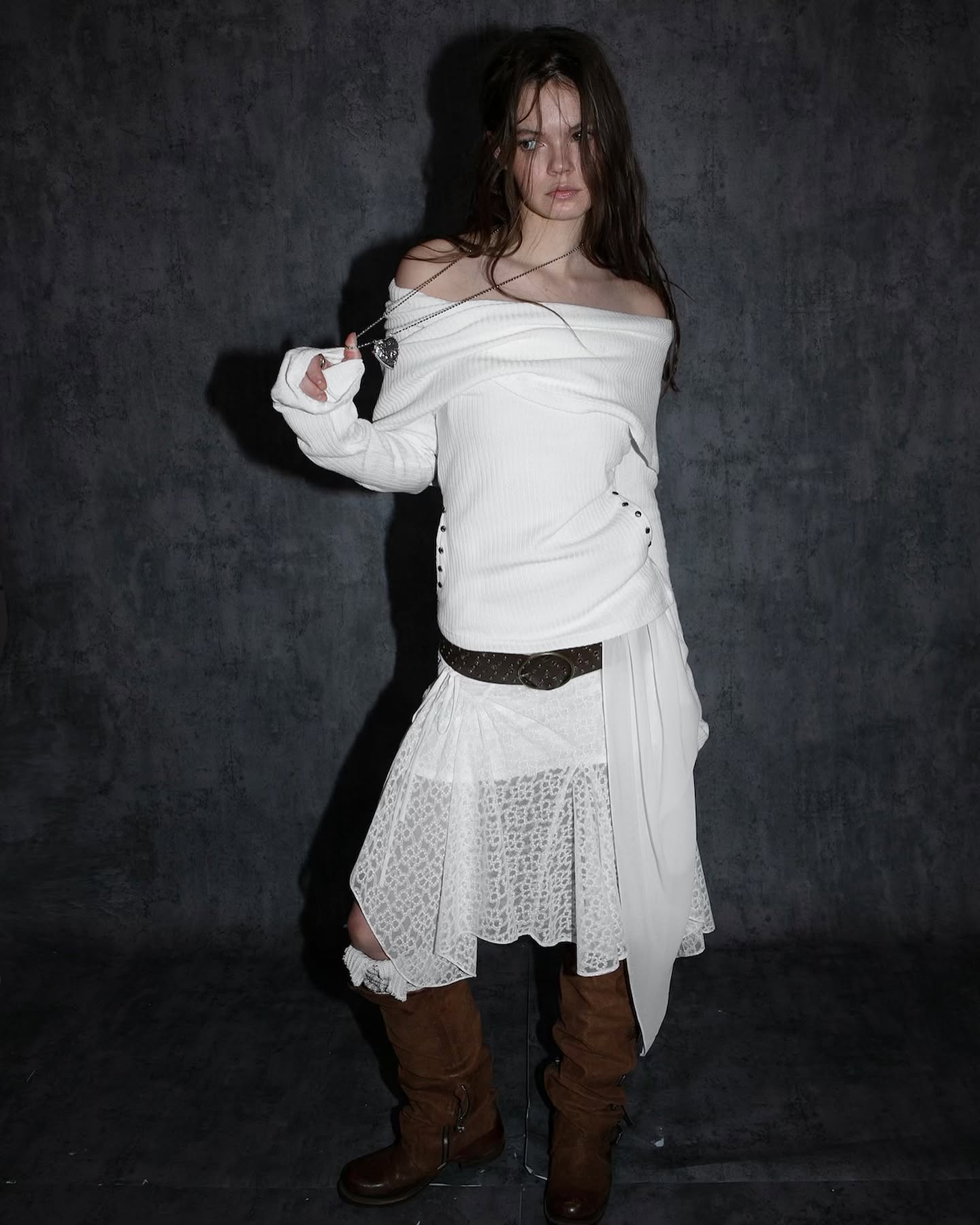 a full shot of a woman in a white offtheshoulder dress with a lace overlay skirt.  she is wearing brown boots and a necklace. the background is a dark grey. the woman is posing for the photo. the overall style is gothic or alternative.