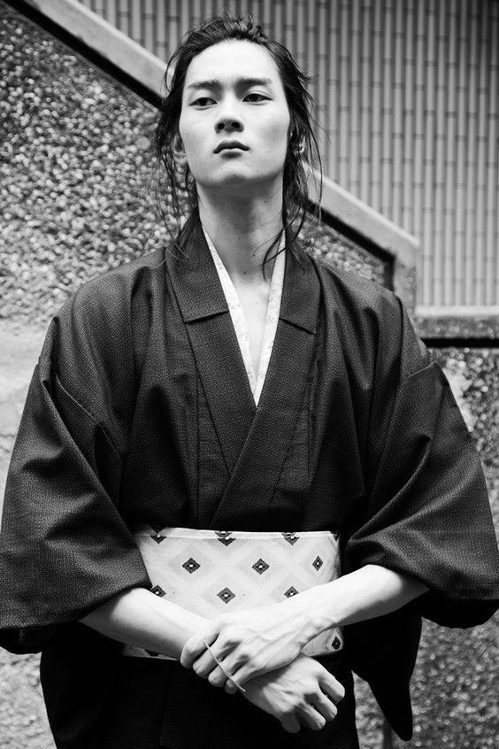 a black and white portrait of a young japanese man with long hair. he is wearing a traditional kimono and is standing in front of a textured wall. the man is looking directly at the camera with a serious expression.  the image has a strong contrast between light and shadow, highlighting the texture of the kimono and the man's hair.