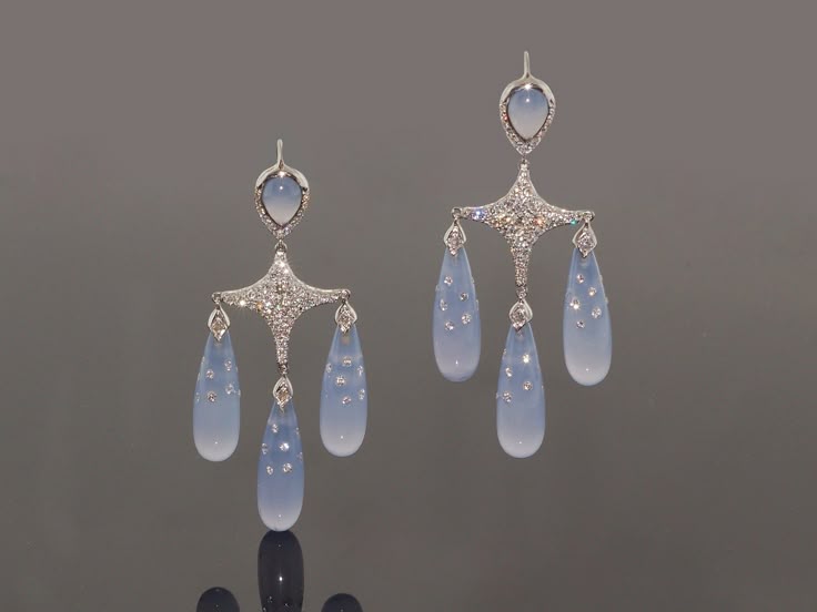 two chandelier earrings are displayed against a grey background. the earrings consist of a teardrop stone at the top, connecting to a diamondencrusted center, from which additional teardrop stones hang. the teardrop stones are a pale blue color and adorned with tiny gems. the metal parts of the earrings appear to be white gold and are heavily inlaid with diamonds.