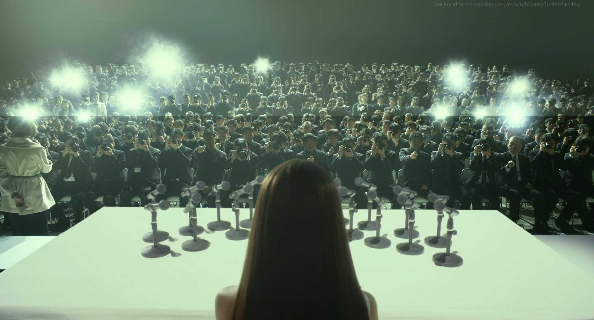 a woman stands on stage in front of a large crowd of people. the crowd is all dressed in black and they are holding cameras. the woman is facing the camera with her back to the crowd. there are a few microphones on the stage in front of her and many lights are flashing. it looks like a press conference or an important event. the scene is lit in a way that makes the crowd look shadowy, highlighting the woman and the microphones on the stage. the image has a feeling of mystery and anticipation.