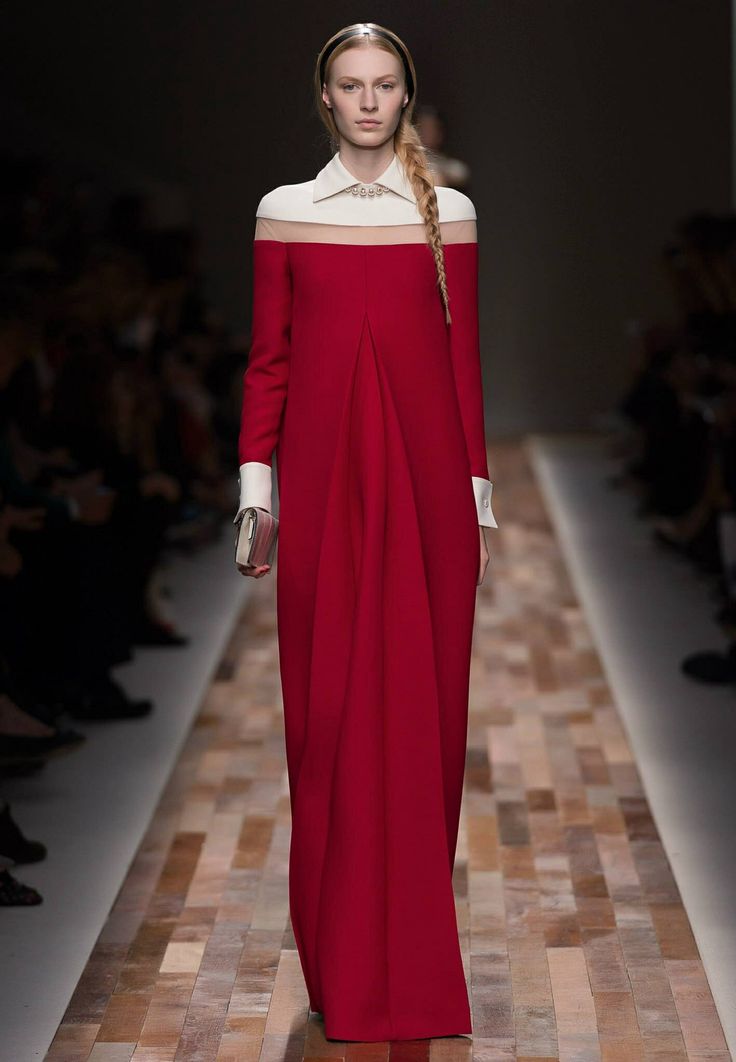 a blonde model walks the runway in a long red dress with a white collar and cuffs. the dress has a simple design with a slight flare at the bottom. she has a white clutch and her blonde hair is styled in a braid. the model is walking on a wooden floor. the background is blurred.