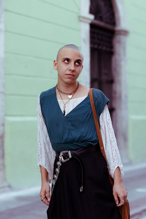 a closeup shot of a young woman with a shaved head, wearing a teal top with lace sleeves, a black skirt, and a brown purse. she's adorned with necklaces and a silver belt chain. the backdrop is a city street with a green building and an old doorway. the woman is looking upward, possibly at something offcamera.  the overall impression is one of fashion and style, with a slightly bohemian or alternative aesthetic.