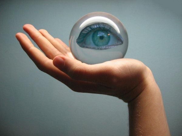 a hand holds a clear crystal ball which contains an eye. the eye is turquoise, with visible eyelashes. the hand is fairskinned, with fingers extended and slightly curved. the arm is visible, extending from the hand. the background is a soft bluegray.