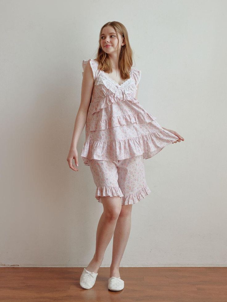 a young woman is standing against a lightcolored wall. she's wearing a pretty pink pajama set. the set consists of a sleeveless top with ruffled tiers and a delicate white lace trim at the neckline, and matching ruffled shorts. the fabric features a small, delicate floral print.  she is wearing white slippers and is posing slightly sideways, looking off to her left. the background is simple, allowing the outfit to be the focus. the overall image is bright and airy.