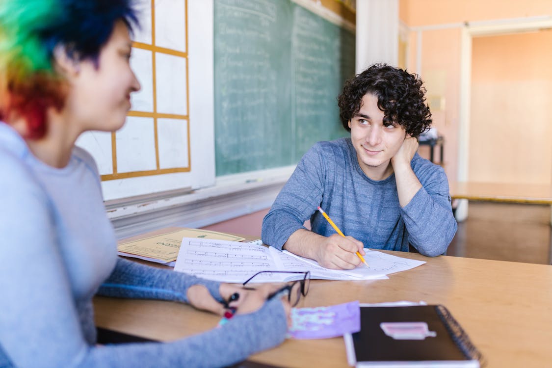two students are sitting at a desk in a classroom. one student has curly hair and is holding a pencil, looking at the other student with a smile. the other student has colorful hair. there are books, notebooks, sheet music, and glasses on the desk.