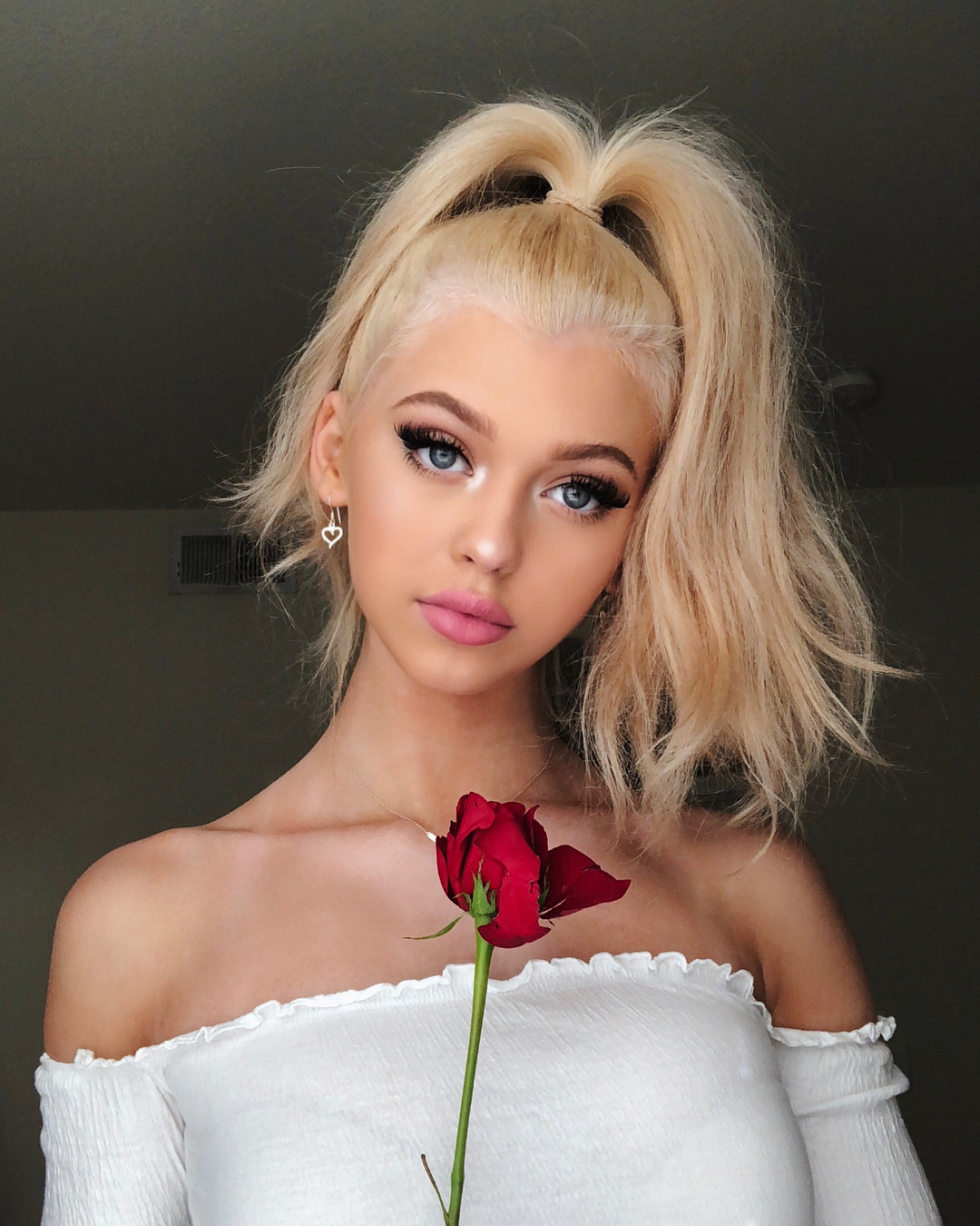 a woman with blonde hair in a high ponytail holds a red rose in front of her. she is wearing a white offtheshoulder top and heartshaped earrings. she has blue eyes and is wearing makeup, including mascara and lipstick.