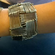a closeup image of a woman's arm wearing a bracelet made from safety pins. the bracelet is a unique and stylish design, made with multiple safety pins woven together to create a geometric pattern. the bracelet is silver in color and is worn on the woman's left wrist. the background is blurred, focusing attention on the bracelet.