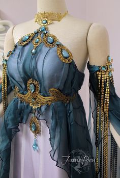 a closeup shot showcases a stunning teal and white gown with intricate gold and turquoise embellishments. the dress, displayed on a mannequin, features a halter neckline, a flowing skirt, and delicate, beaded sleeves. a thick gold belt with a central turquoise stone accentuates the waist. the overall design evokes a sense of ethereal beauty and fantasy, reminiscent of a goddess or elven attire.
