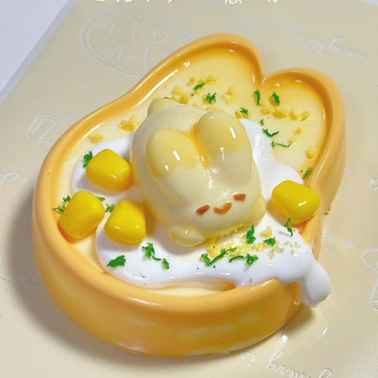 the image shows a cute bunnyshaped dessert. the dessert is in a heartshaped bowl filled with yellow pudding. a bunnyshaped confection rests on top of a dollop of whipped cream, surrounded by pieces of corn and green garnish. the overall aesthetic is very cute and appealing.