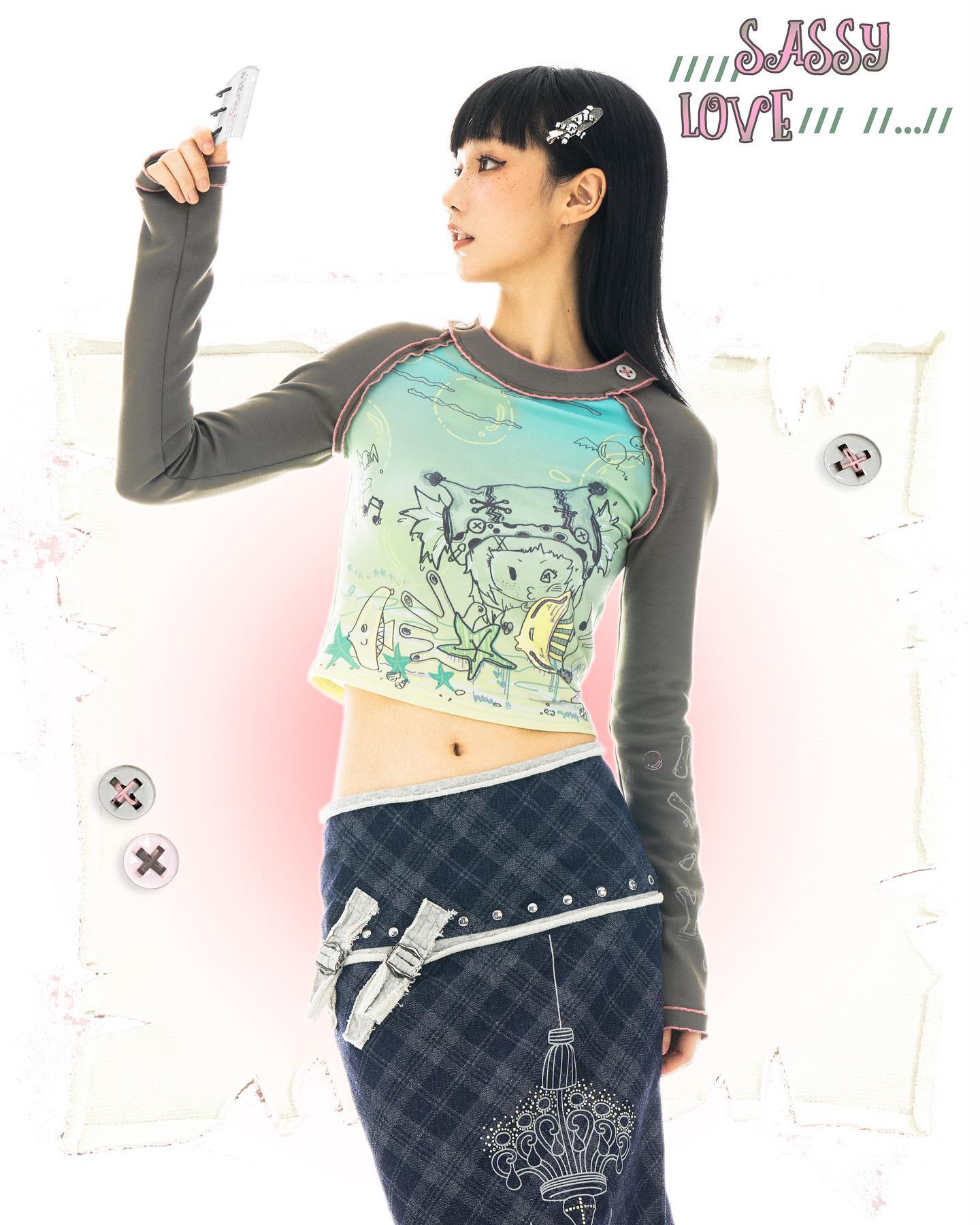 the image features a young asian woman modeling a trendy outfit. she's wearing a longsleeved, pastelcolored graphic tee with a whimsical design, paired with a dark plaid denim skirt adorned with silver studs and decorative fabric accents. her hair is styled with black hair clips, and she's holding a small object in her hand. the background is a light pink and offwhite with stylized design elements. the overall style is a blend of kawaii, harajuku, and alternative fashion aesthetics.  the words "sassy love" are overlaid on the image.