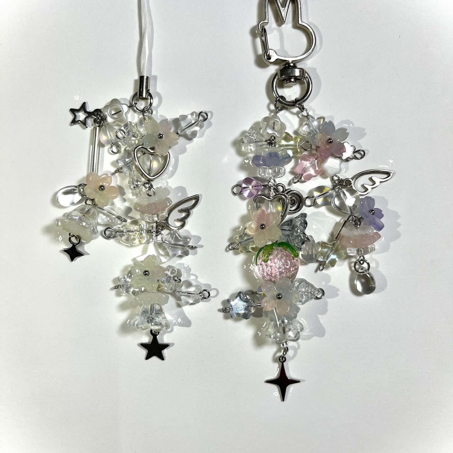 two handmade beaded keychains are displayed on a white surface. both keychains feature a combination of clear and pastelcolored flower beads, clear beads, and silver hardware. one keychain has a star charm, heart charm, and wings, and is attached to a white cord. the other keychain has a strawberry bead, wings, and a star charm, and is attached to a silver bunnyshaped clip.