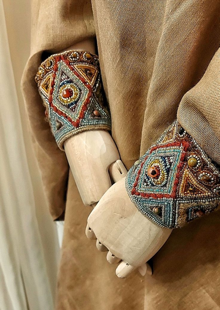a close up image of a garment with intricate beaded embroidery detail on the cuffs. the cuffs are on the wrists of a wooden mannequin hand. the fabric is a light brown colour and the embroidery is colorful and geometric. the image highlights the detail and craftsmanship of the garment.