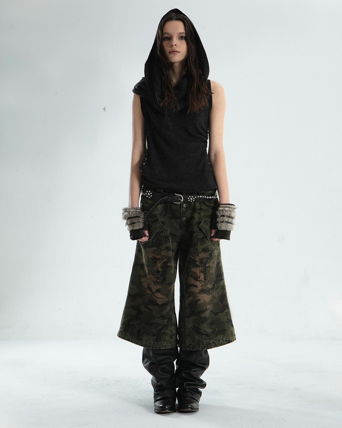 the image shows a female fashion model standing against a plain white background. she's wearing a dark gray sleeveless cowl neck hooded top,  a studded belt, camouflage wideleg cropped pants, and black boots that are slightly folded at the ankle.  she also sports gray fingerless gloves. the overall style is grunge or alternative, with a focus on urban and slightly edgy elements.  the photo is a fullbody shot taken in a studio setting.