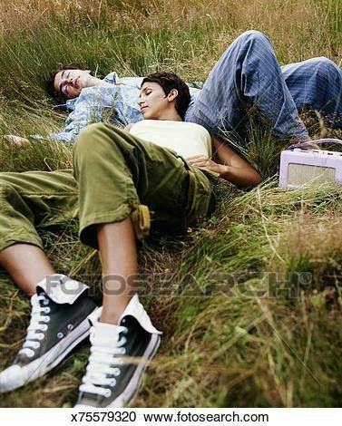 two people lay in the grass relaxing and enjoying the day.  the female has her arm around the male.  they are listening to music from their portable radio.content: serenepeople: man, womanenvironment: outdoors, field, grasslighting: bright, natural light, daytimeperspective: high anglekw: grass, couple, relaxation, leisure, music, radio, nature, tranquility, summer, peace, love, togetherness, enjoyment, outdoors, field, meadow, green, blue jeans, tshirt, sneakers, portable radio, vintage, carefree, happy, content