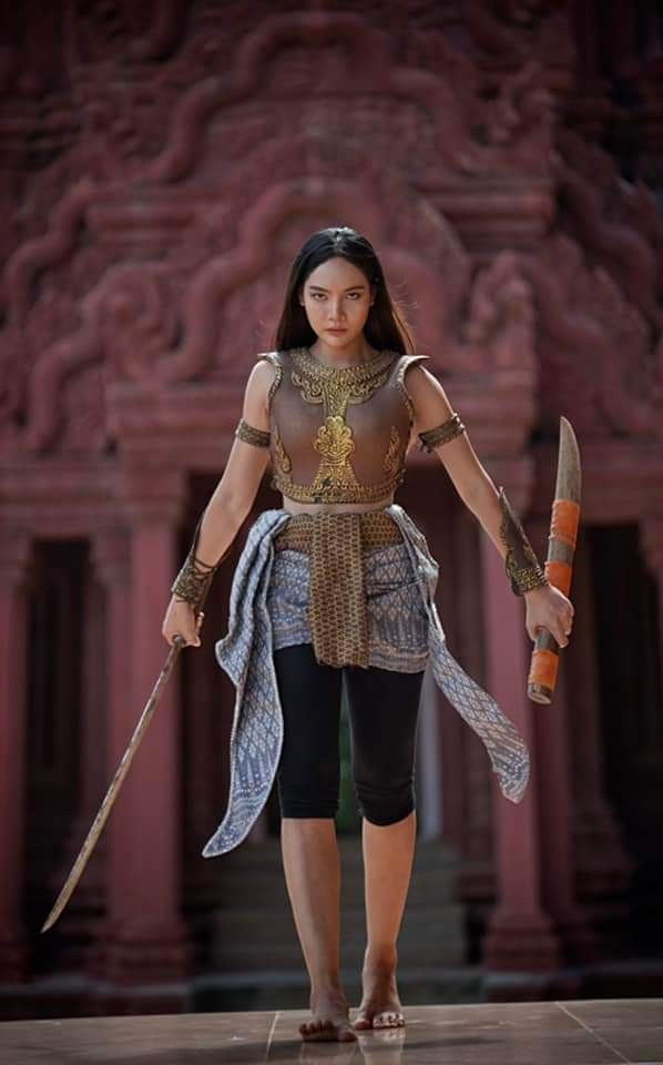 a captivating image of a young asian woman, possibly thai, dressed as a female warrior. she stands confidently in front of an ancient red temple, showcasing her traditional attire. her outfit features elaborate armor, a patterned wrap, and black leggings.  she holds two swords, one a long sword and the other a shorter, possibly ceremonial blade. the temple's intricate carvings form a dramatic backdrop, enhancing the overall powerful and historical feel of the image. the woman's expression is serious and determined, reinforcing her image as a formidable warrior. her barefoot stance adds to the grounding and strength of the composition.