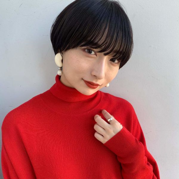 a young woman with short black hair, wearing a red turtleneck sweater, smiles at the camera. she is wearing earrings and a ring. her hair is styled in a bob with a fringe. she has a natural and fresh look, with light makeup and a friendly expression.