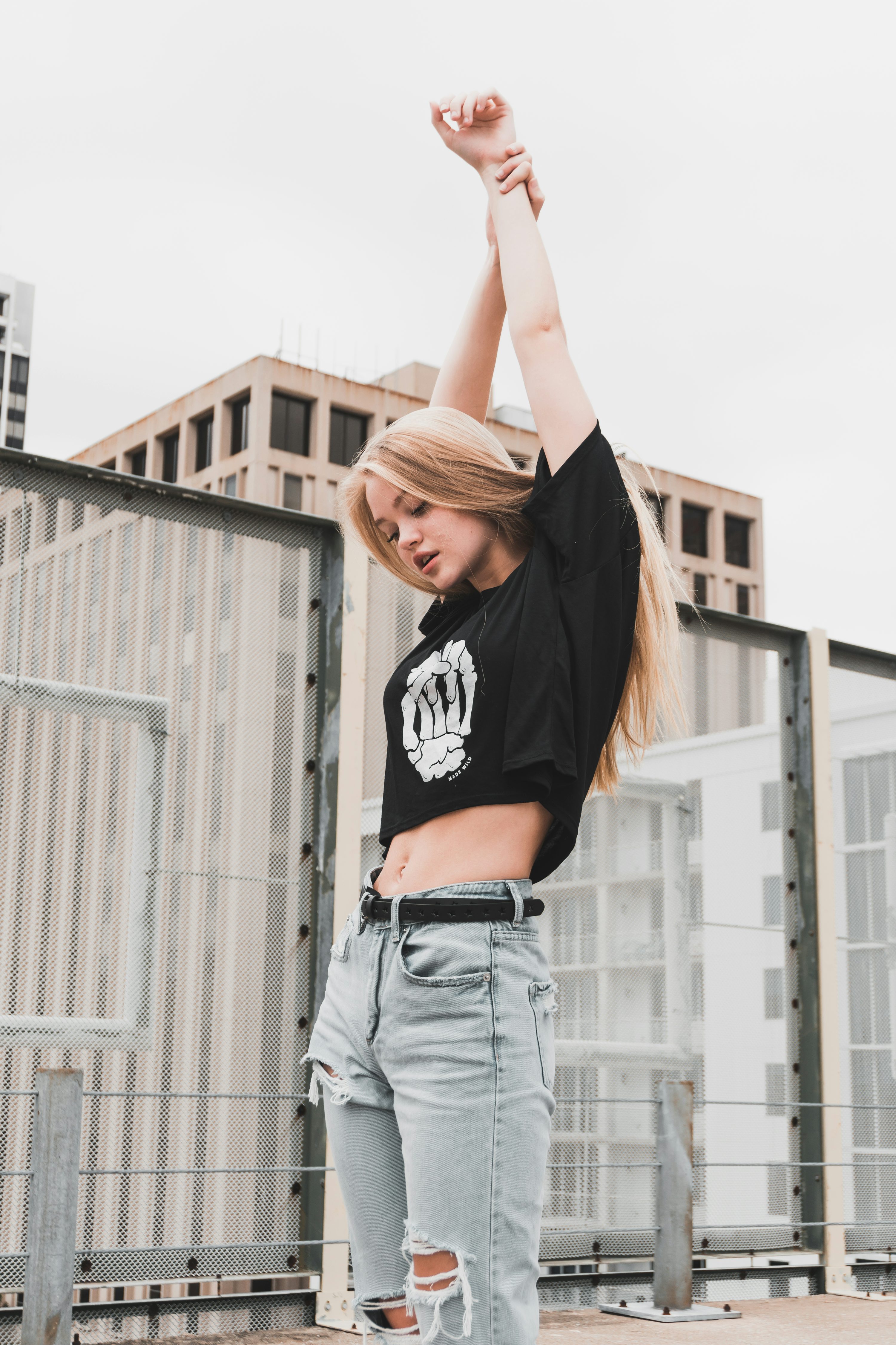 a young blonde woman is standing outdoors in a city setting, posing with her arms raised above her head. she is wearing a black crop top with a skeleton hands design, light blue ripped jeans, and a black belt. the background includes modern buildings and a metal fence. the overall style is casual and urban.  she appears happy and confident.