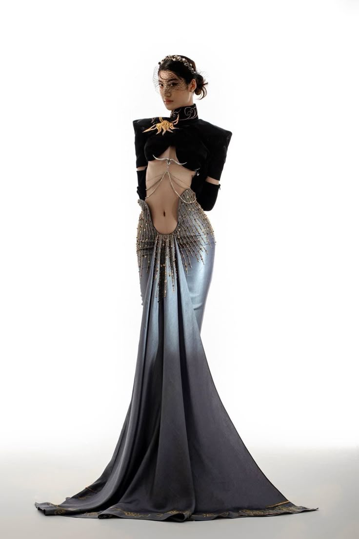 a fulllength studio shot captures a woman modeling a striking and elegant gown. the dress features a black highnecked top with padded shoulders and a gold applique, transitioning into a silver, beaded midsection with an exposed midriff. the bottom of the gown is a silver and grey flowing skirt with a train, trimmed with gold accents. she is also wearing a headpiece with a sheer veil. the model is standing against a bright white backdrop.