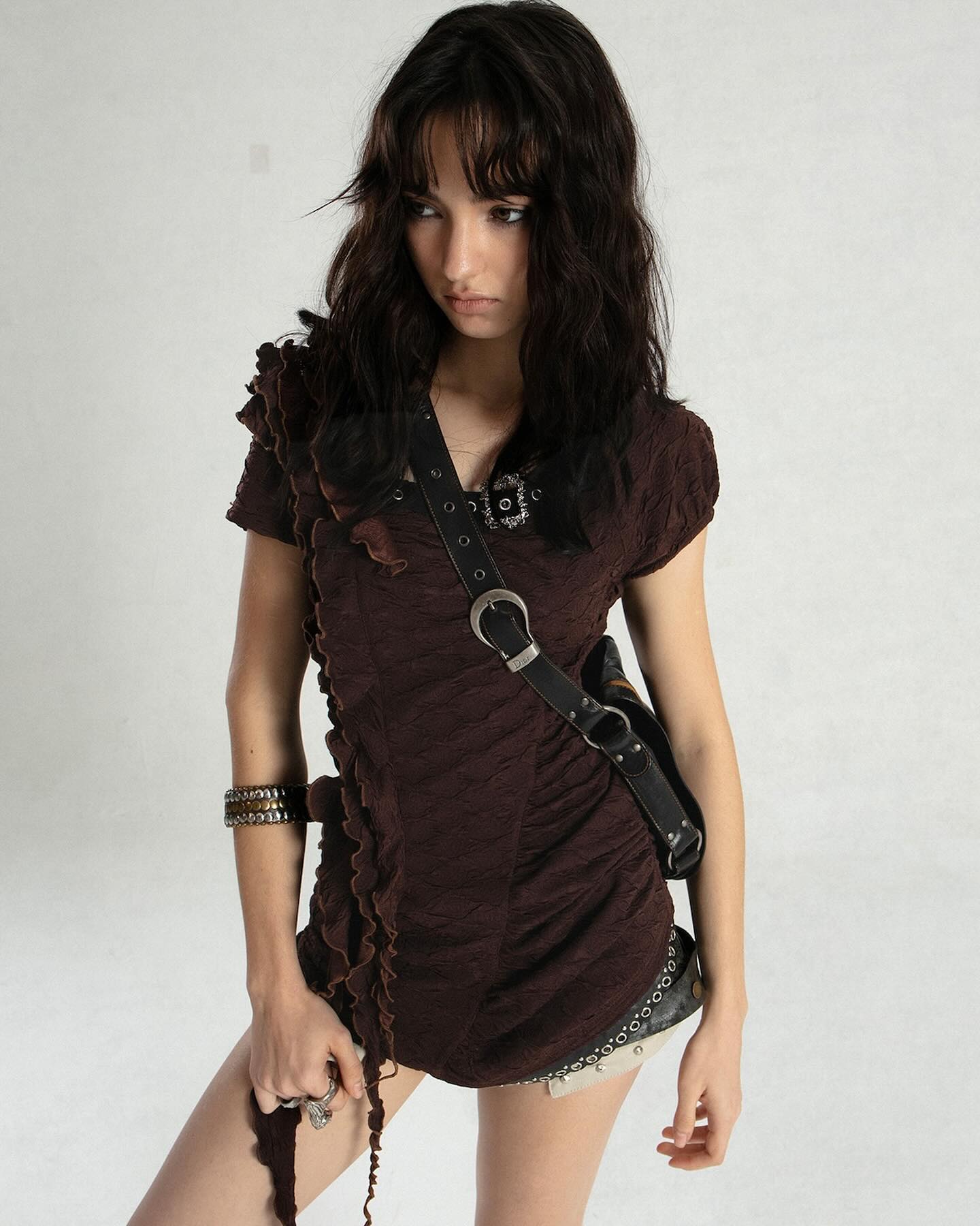 a young woman with long, dark brown hair and bangs is captured in a studio shot against a plain white backdrop. she is wearing a brown, ruffled, textured mini dress with metal accents and a leather crossbody bag. her accessories include bracelets, a necklace, and rings. her expression is somewhat serious and pensive as she gazes downward. the overall image portrays a fashionforward, casual style.