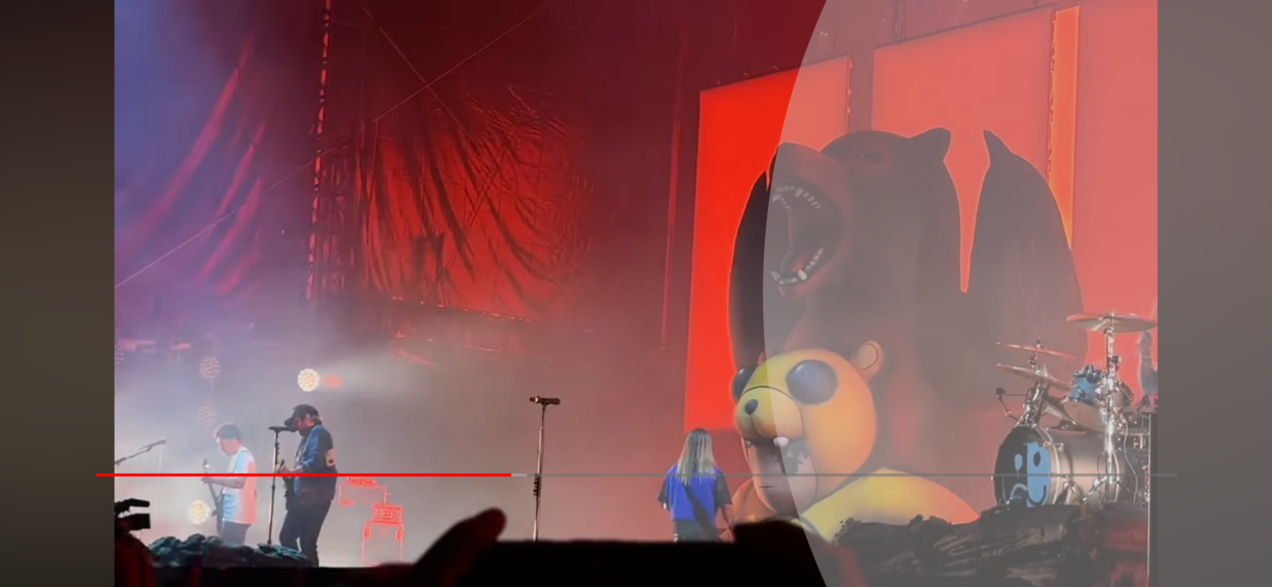 a live music performance in a concert setting. the stage is dimly lit with a spotlight illuminating the band in front of a large red screen. the band consists of three members: a guitarist, a vocalist, and a drummer. the guitarist and the vocalist are playing their instruments in front of a large screen with a cartoon of a bear and a bear cub. the drummer is playing a drumset in front of the screen. the crowd is visible in the background, though not in focus.