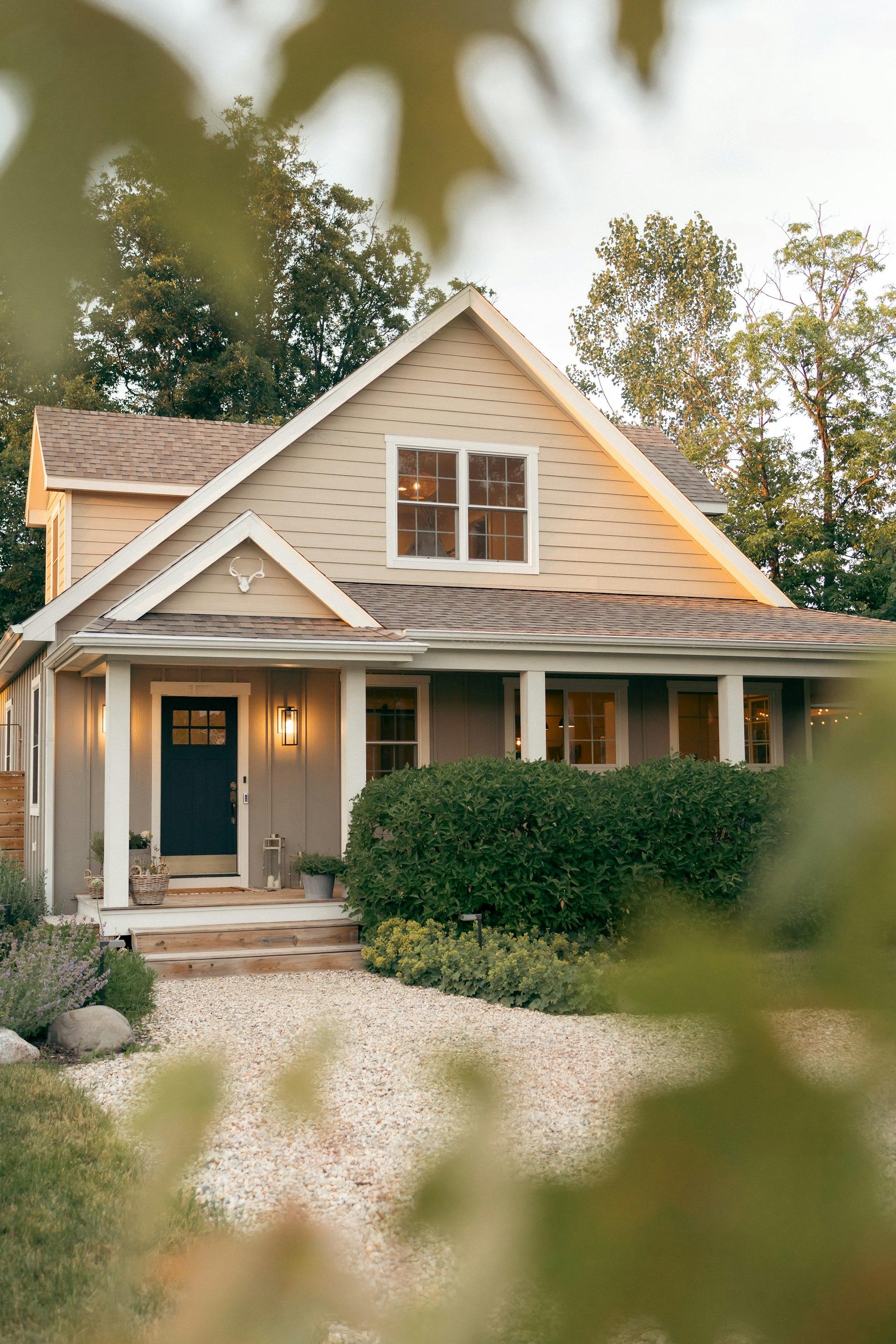 the image shows a beige twostory house with a gabled roof, white columns on the porch, and a dark blue front door. the house is surrounded by wellmaintained landscaping, including a neatly trimmed hedge, shrubs, and a gravel driveway.  the scene is viewed slightly from an angle through leafy branches in the foreground, and the lighting suggests it's either late afternoon or early evening.  the overall style of the house suggests a blend of craftsman and farmhouse architectural elements.