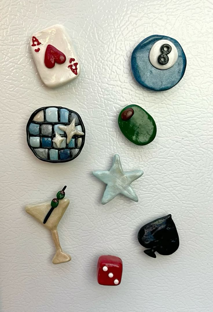 this image contains various magnets. there is an ace of hearts, an 8ball, a disco ball with two stars on it, an olive, a star, a martini, a spade, and a dice.