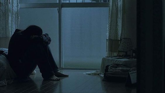 a person sits on the floor in the fetal position, their back to the camera, in a dimly lit room. the figure appears to be illuminated by the light coming in from a large window. the mood is somber and suggestive of sadness or despair.