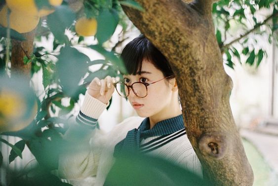 a young woman with short dark hair and glasses is partially hidden behind an orange tree. she's wearing a white sweater with navy blue stripes, and her gaze is directed away from the camera.  the natural lighting of the outdoor setting illuminates the scene.