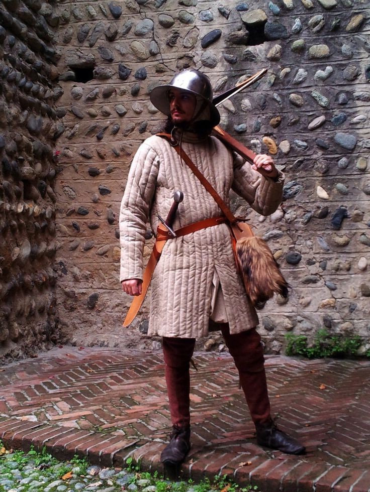 the image shows a man dressed in a meticulously detailed 15thcentury medieval soldier's costume. he's wearing a padded aketon (quilted jacket), hose (leggings), leather belt with a dagger and sword, and a fur pouch. he carries a crossbow (or possibly a longbow) over his shoulder. the helmet is a simple, closefitting type typical of the era. the backdrop is a roughhewn stone wall and a brick pavement. the overall impression is that of a historical reenactor, carefully portraying a soldier from the late medieval period.