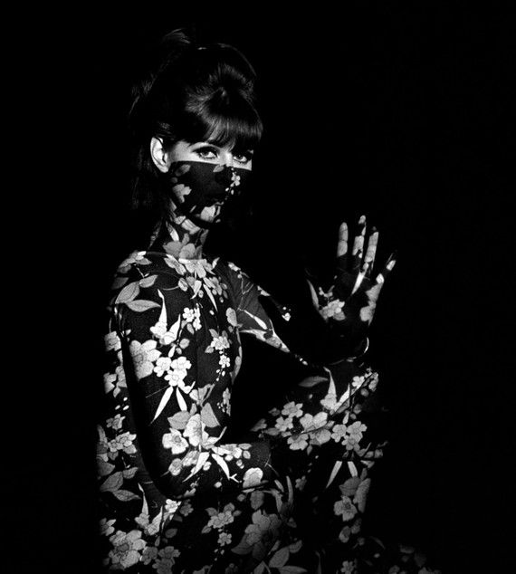 the image is a striking black and white portrait of a female model from the 1960s. she is wearing a formfitting, floralprint bodysuit that covers her entire body, except for her hair and a portion of her face above a dark mask.  her hair is styled in a classic 60s bouffant. one hand is raised in a gesture that adds to the enigmatic nature of the photograph. the dark background emphasizes the model and her outfit. the overall effect is one of mystery and high fashion, indicative of the artistic photography styles of that era. the floral pattern is crisp against the dark background and the contrast between the black and white enhances the visual impact.