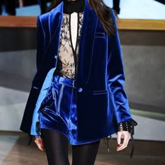 a woman with long dark hair, wearing a blue velvet blazer and matching shorts, poses for a fashion shoot. her outfit is completed with a delicate lace top and sleek black tights. she looks confident and stylish, showcasing the latest trends in fashion.
