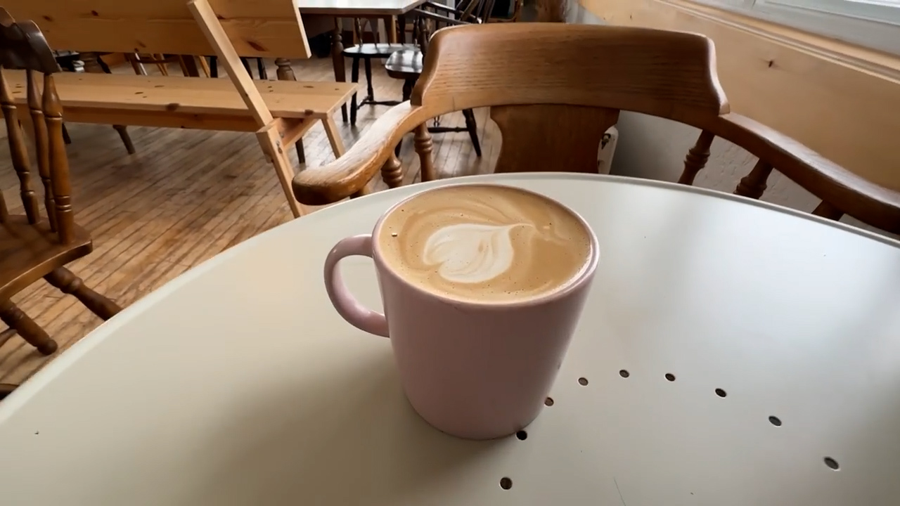 a pink mug of coffee with latte art sits on a white table in a cafe. the background shows wooden chairs and tables.