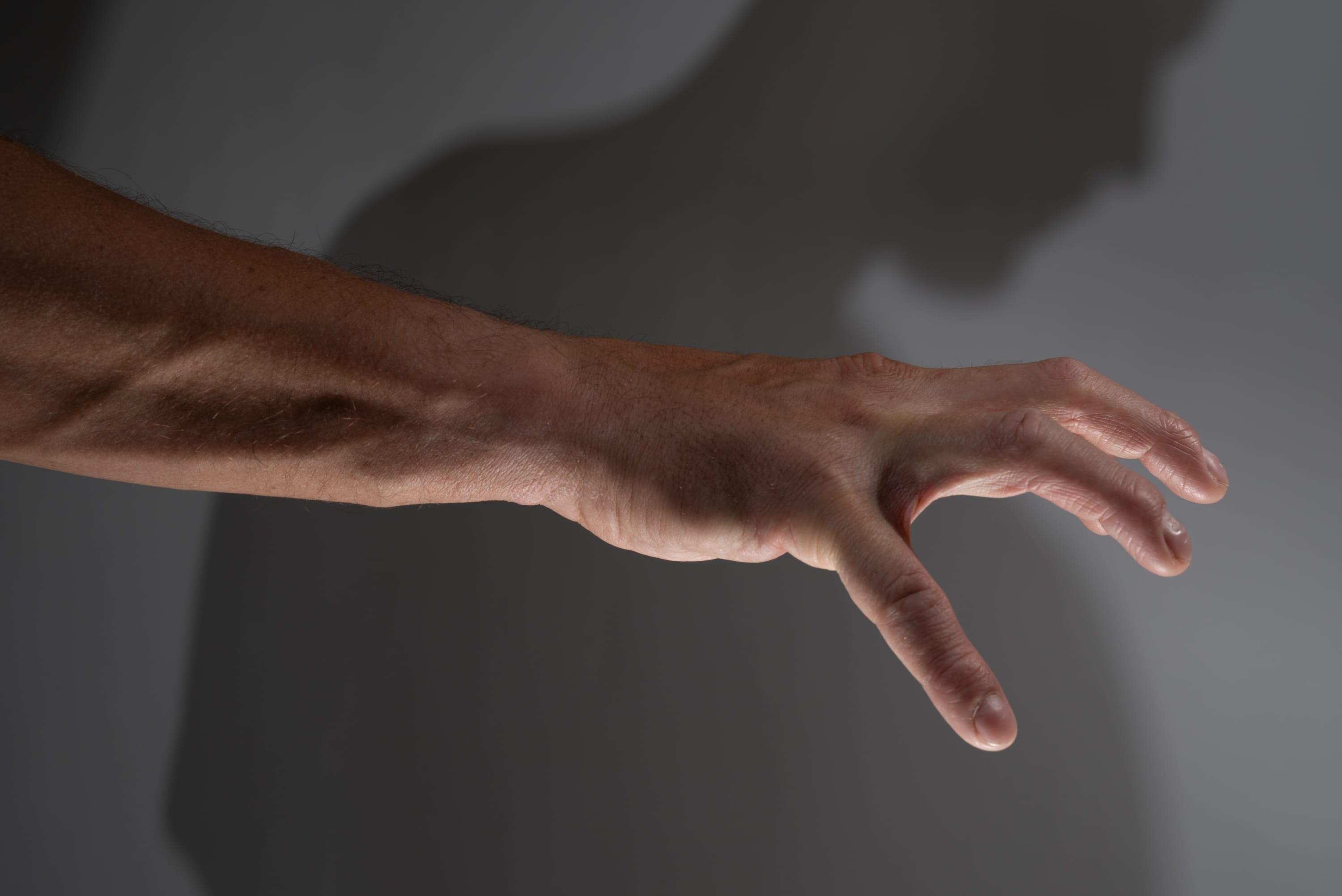 a man's arm extends from the left side of the frame, fingers splayed open. his shadow is cast on the grey background. the image uses studio lighting.
