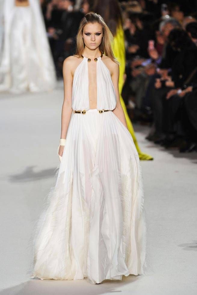 a beautiful woman in a white flowing dress with a gold belt is walking down the runway at a fashion show. she is smiling and posing for the cameras, looking absolutely stunning. the dress is very elegant and stylish, and it perfectly complements her beautiful features. she is wearing her hair down in loose waves, and her makeup is soft and natural. the overall look is very sophisticated and glamorous.