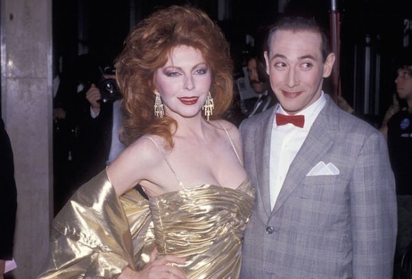 a woman in a gold gown and a man in a gray suit with a red bowtie are smiling for a photo. the man is on the right and the woman is on the left. the woman has long, red hair. the man is wearing a black tie under his bowtie.  they are at a formal event.