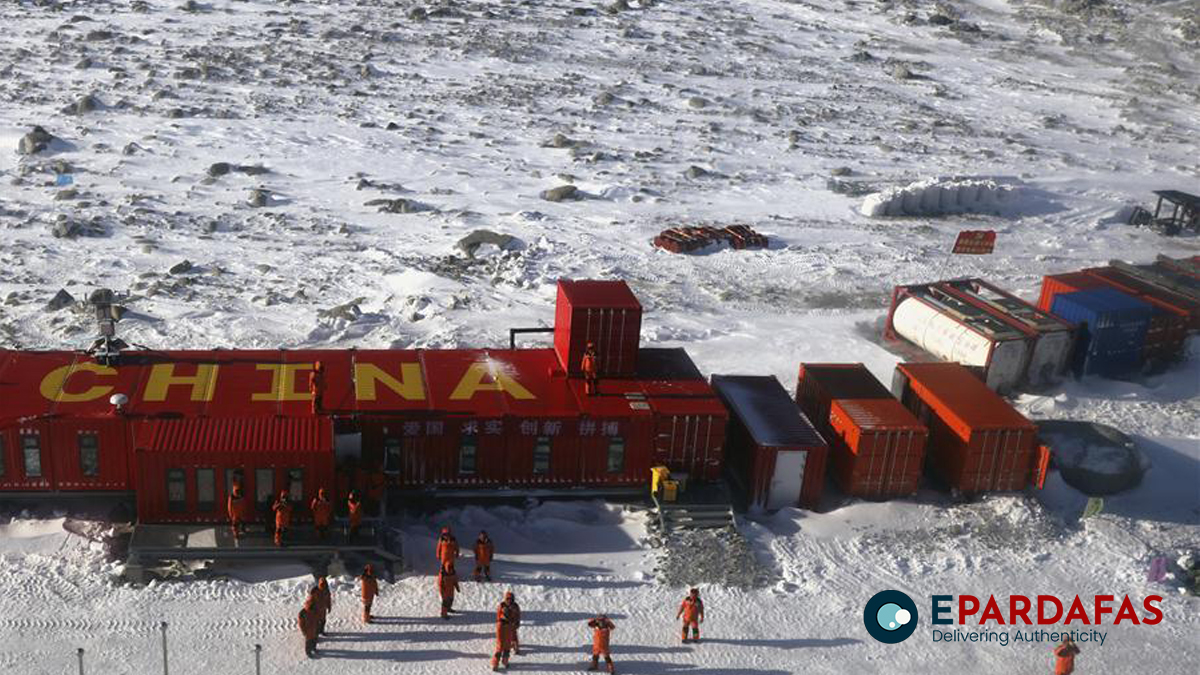 an aerial view of the chinese research station in antarctica. the station is made up of a number of red containers with the word "china" written in yellow letters on the roof. there are a number of workers in red suits standing around the station, and a chinese flag is flying in the wind. the station is surrounded by snow and ice, and the landscape is desolate and remote.