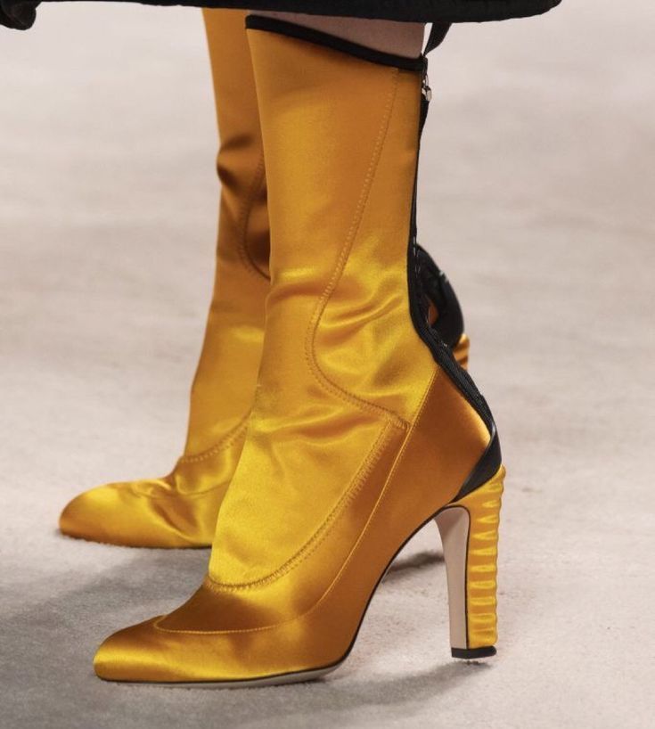 the image shows a pair of yellow satin highheeled boots. the boots have a pointy toe, a ribbed high heel, and a black trim detail at the top and back of the boot. there is a zipper on the back of the boot.