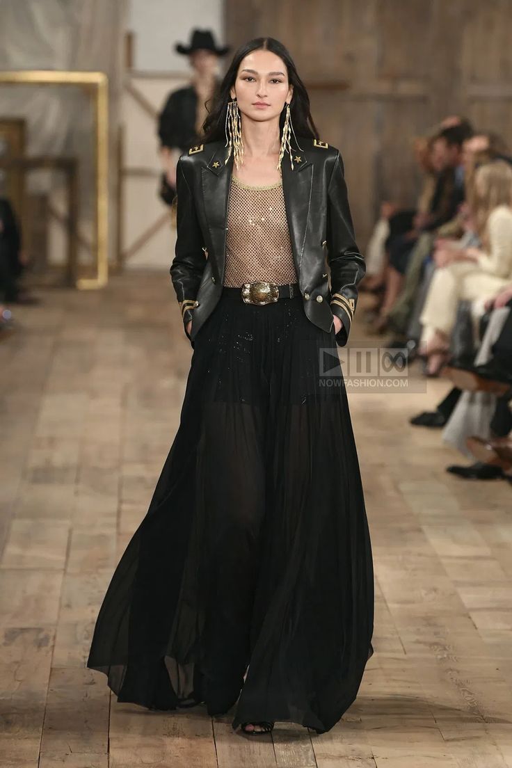 a fashion model walks down a runway wearing a black leather jacket with gold details. the model is also wearing a black sheer dress with a gold belt and long gold earrings. the model is walking down a wooden runway.  there are other people in the background, including a model wearing a cowboy hat.