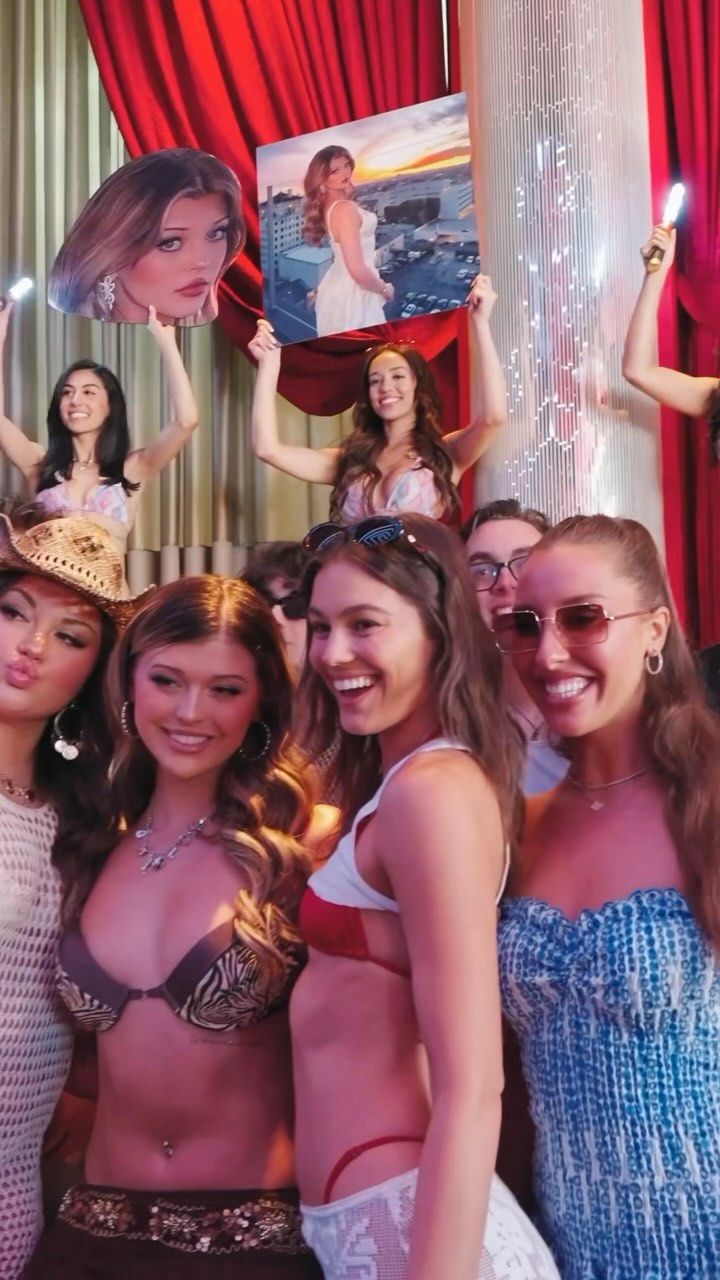 a group of people are posing for a photo at an event or party. there are women in the foreground and in the background, some holding up a cardboard cutout and a poster. the background includes a red curtain and a decorative pillar. the atmosphere appears lively and celebratory.