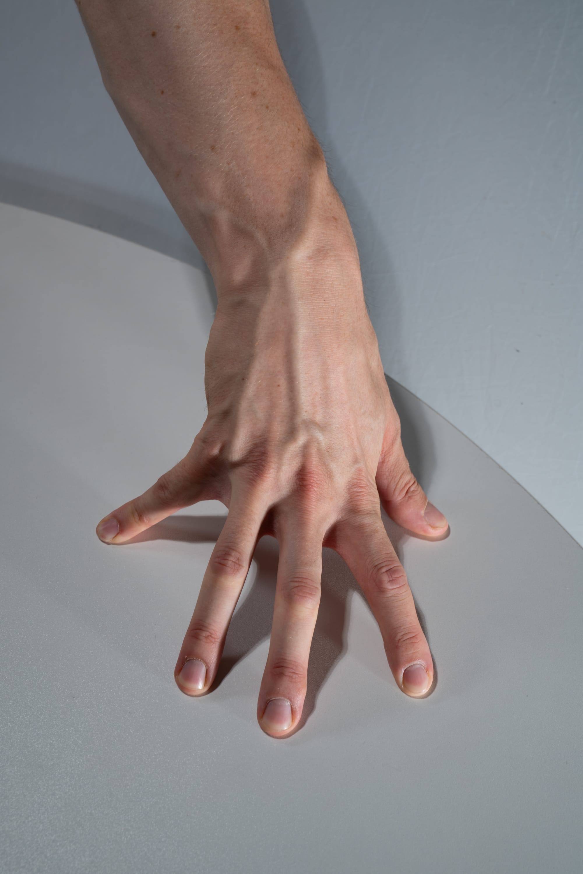 a man's right hand is shown with its fingers spread out on a white surface. the arm is extended and the hand is positioned near the edge of the surface. the hand is illuminated by artificial light, highlighting the veins and details of the skin. the image focuses on the hand and arm, providing a closeup perspective of the subject.