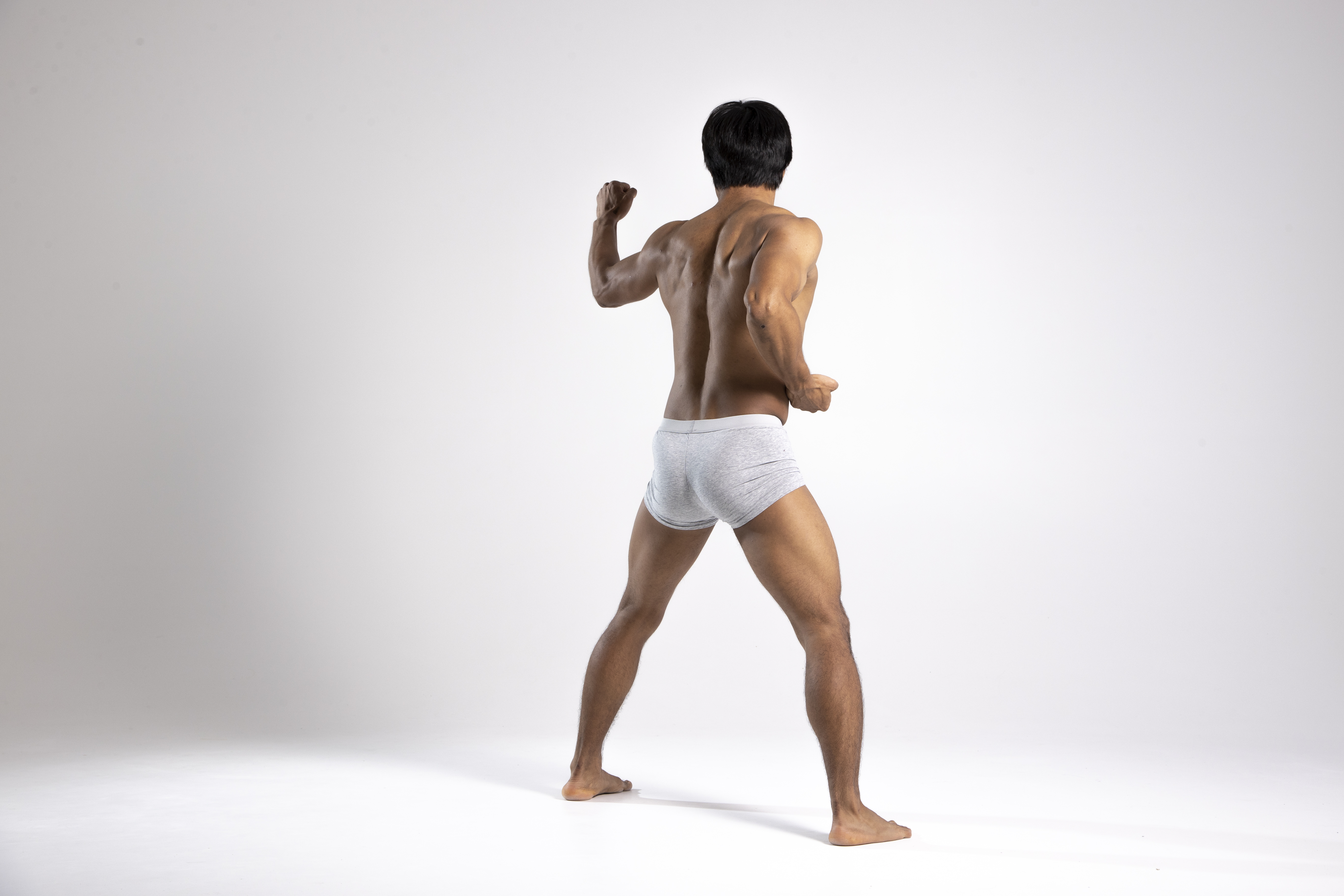 rear view of a muscular, shirtless man wearing underwear, posing in a studio setting. he has black hair and is standing with his back to the camera. his physique suggests he is a bodybuilder or someone with a strong fitness regimen.