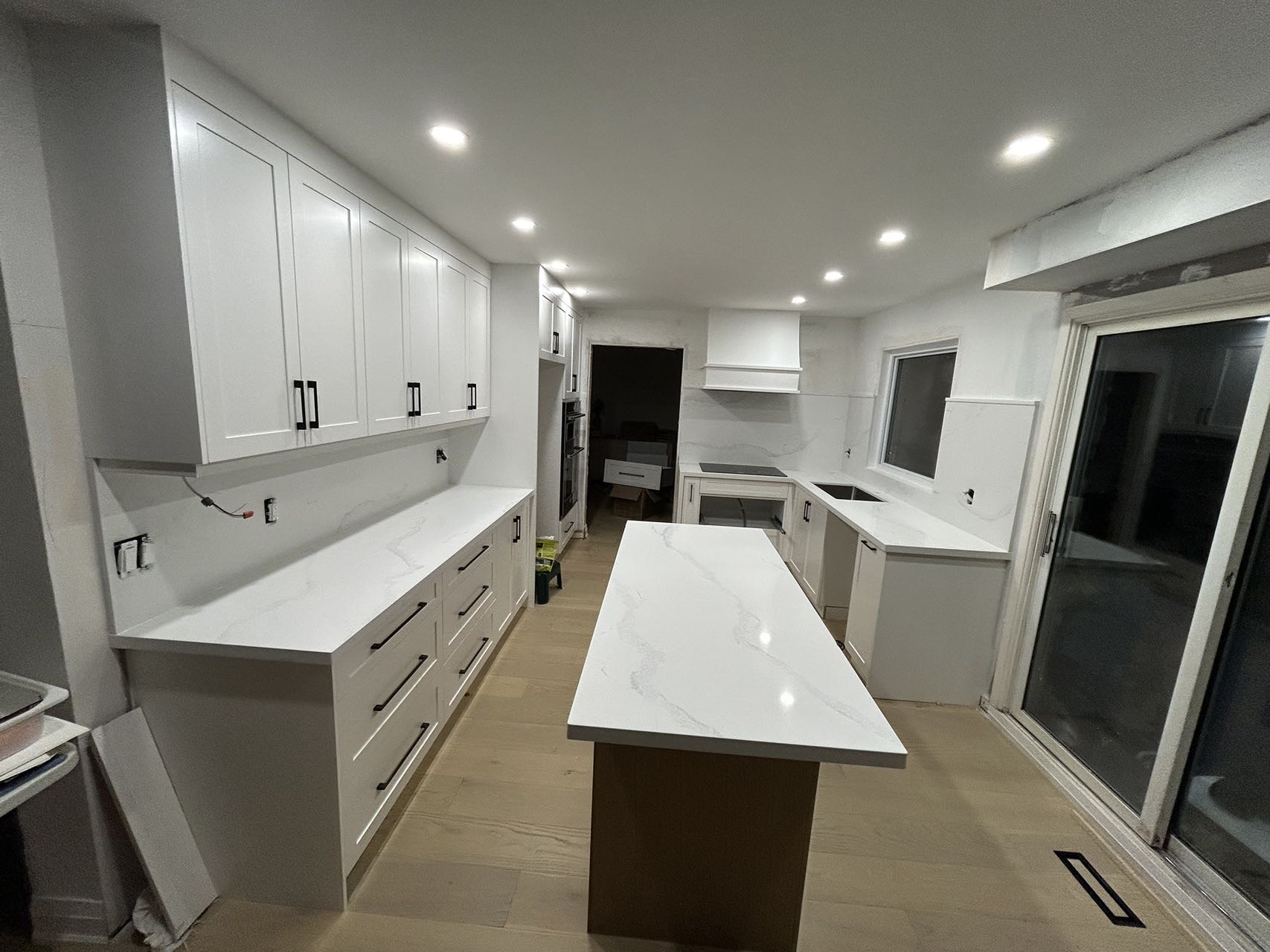 the image shows a newly remodeled kitchen with white shakerstyle cabinets and white quartz countertops.  the kitchen features a large island in the center with the same countertop material. the flooring is lightcolored wood.  the backsplash is white, and recessed lighting illuminates the space.  the cabinets are installed, and some finishing touches are still being completed, including electrical work.  a sliding glass door is visible on the right, leading to the outdoors.  the overall style is modern and clean.
