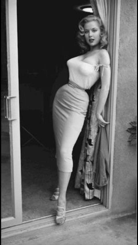 a black and white vintage photograph of a blonde woman standing in a doorway. she is wearing a stylish dress, sandals, and has her hair styled in a classic 1950s pinup look. the image captures a sense of glamour and elegance from a bygone era.