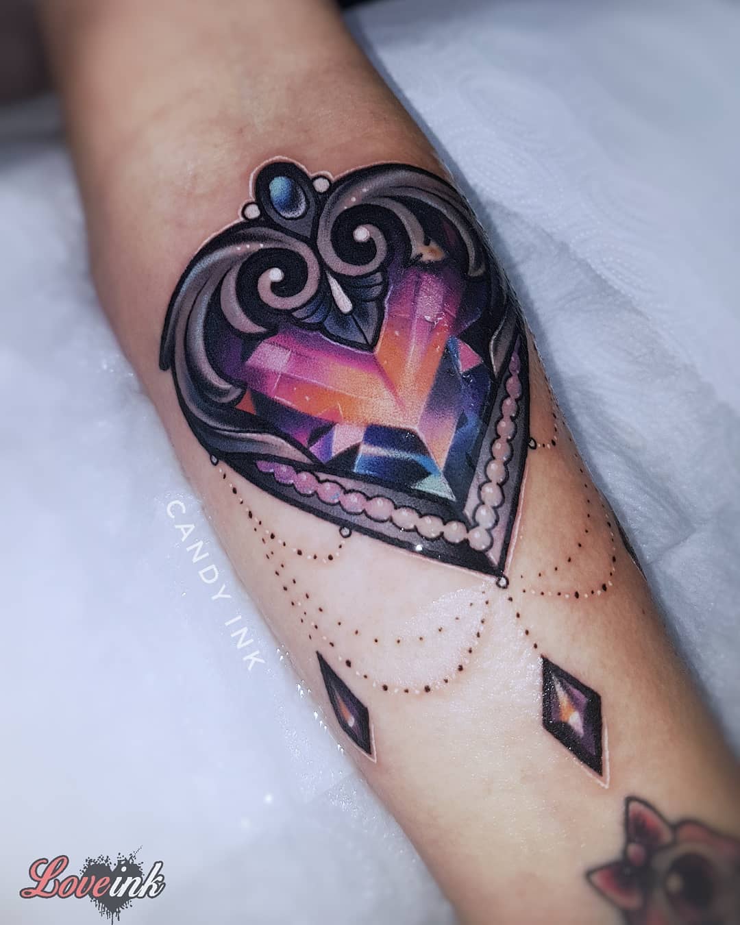 a closeup shot of a neotraditional style tattoo of a heartshaped jewel on someone's forearm. the tattoo has been done by "candy ink" and features hanging jewel accents.  another tattoo by "love ink" is partially visible on the right edge of the image.
