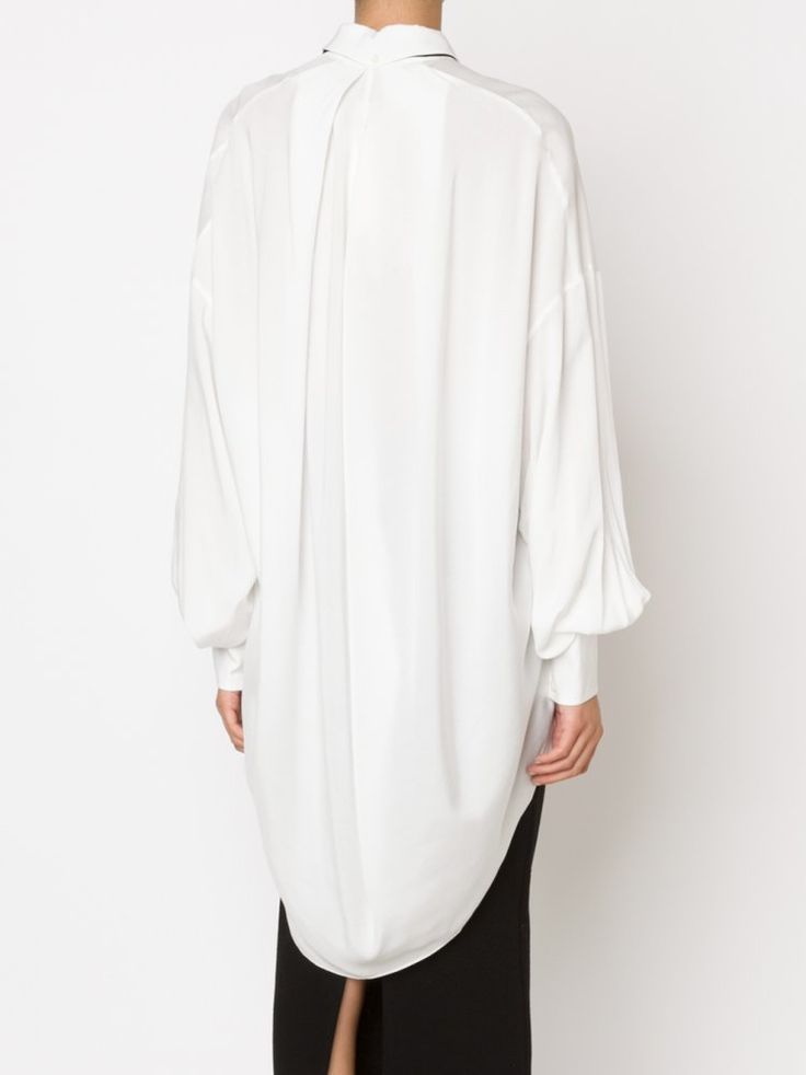 the image shows the back of a person wearing a longsleeved white blouse or shirt. the shirt has a loose, oversized fit with pleats in the back and a collar. the person is also wearing a black skirt or dress that is visible below the hem of the shirt. the background is plain white.